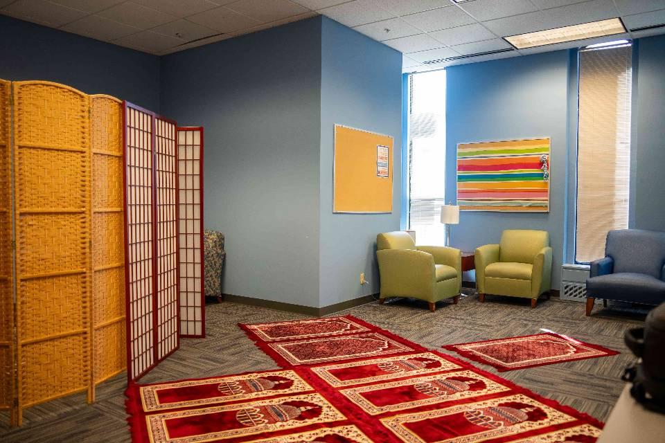 A room with blue walls and comfortable chairs. Prayer rugs are seen lined up on the floor.