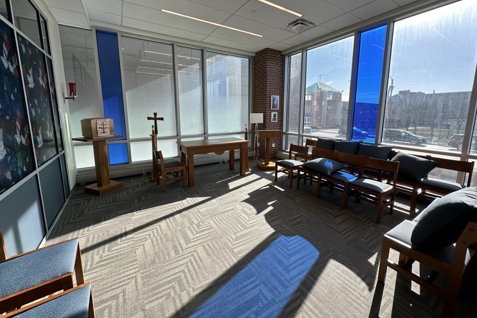 Interior view of a room with padded chairs lining the walls, an altar table, cross, tabernacle and candle holders. There are large windows along along the wall with two stained blue. Sunlight is streaming in, and the sunlight is reflected on the carpet.