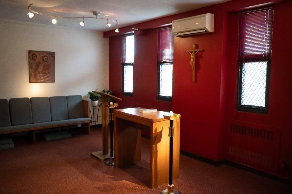 Interior view of a room with an altar, crucifix lectern and candle holder at the front and center, with a padded pew along one wall. A religious painting is visible.