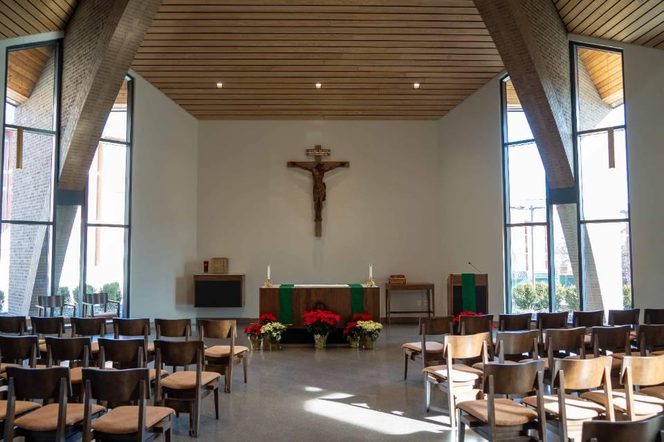 View of a chapel with rows of chairs seen from the back, with an aisle down the middle. At the front, there is an altar, crucifix. Red and white poinsettias are on display. There are tall windows on each side, with a slatted wooden ceiling.