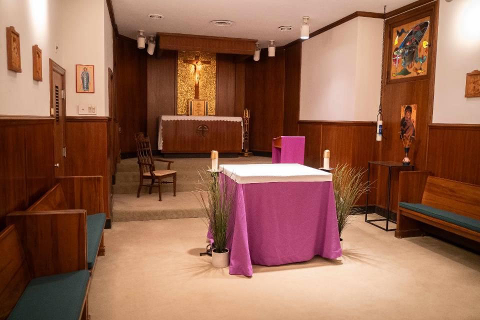 A room with church pews along each wall. There's an altar table in the center. In the background, behind the altar table, there is a long cabinet with a tabernacle sitting on it, with a crucifix on the wall, surrounded by gold wall decoration.. Framed religious artworks are visible on the walls.