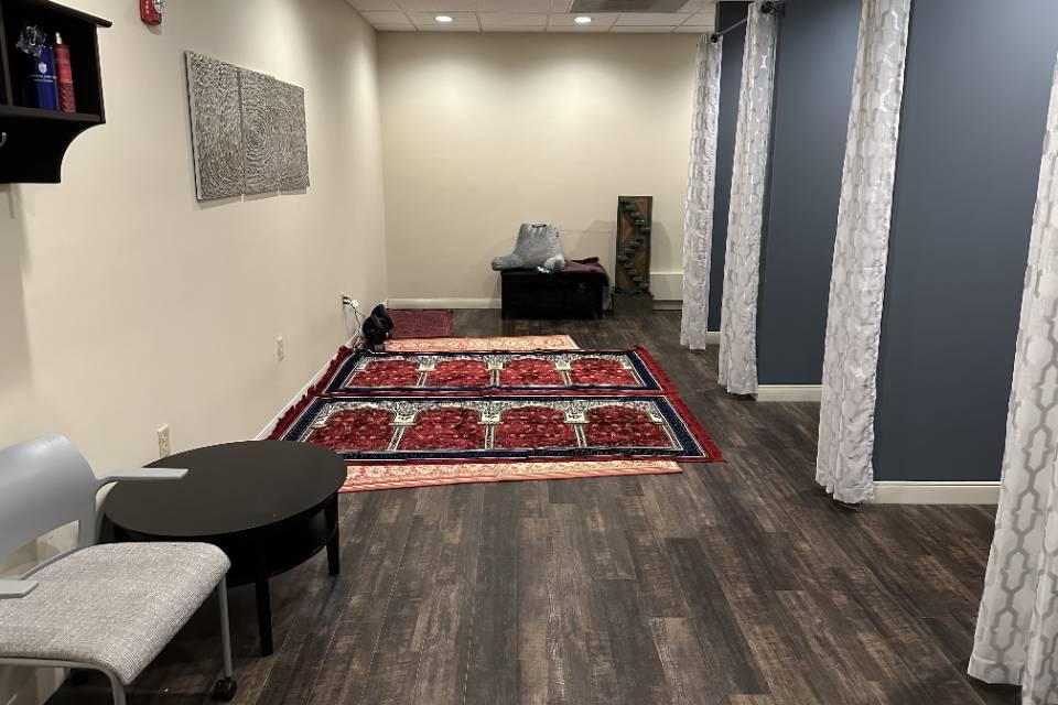 A long narrow room with alcoves on each side with curtains. There are prayer rugs lined up on the floor, as well as some chairs and cushions.